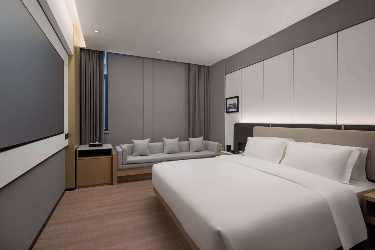 A modern hotel room with a large bed, a sofa, a desk, and a wall-mounted TV.