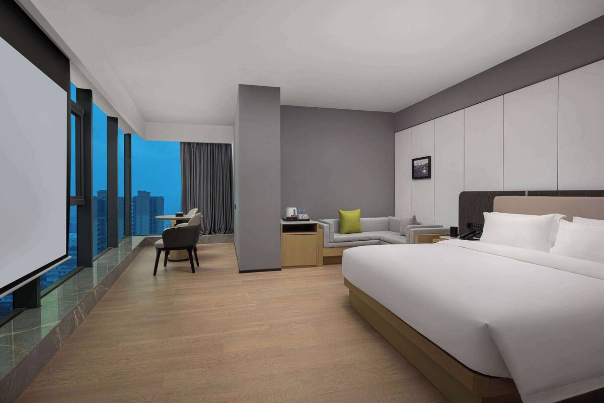 A modern hotel room with a large bed, a desk, a sofa, and a view of the cityscape.