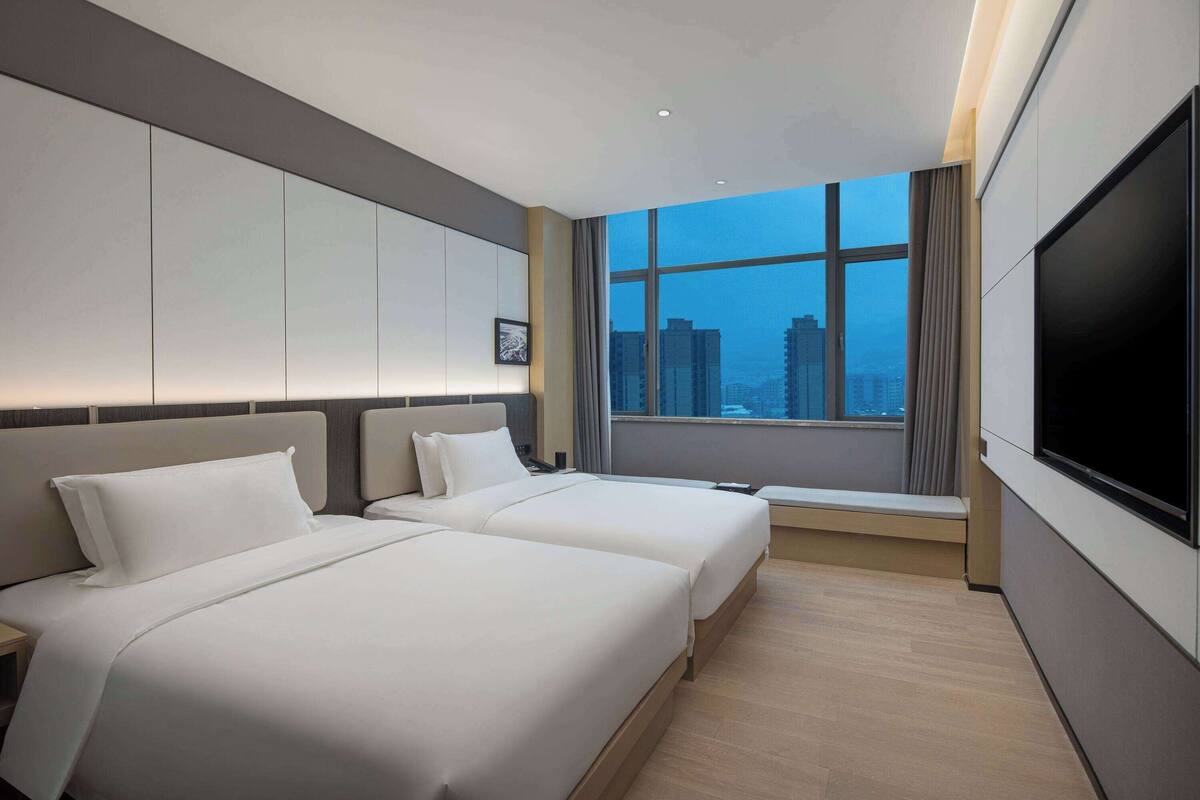 A hotel room with two beds, a large window with a city view, a flat-screen TV, and a bedside table.