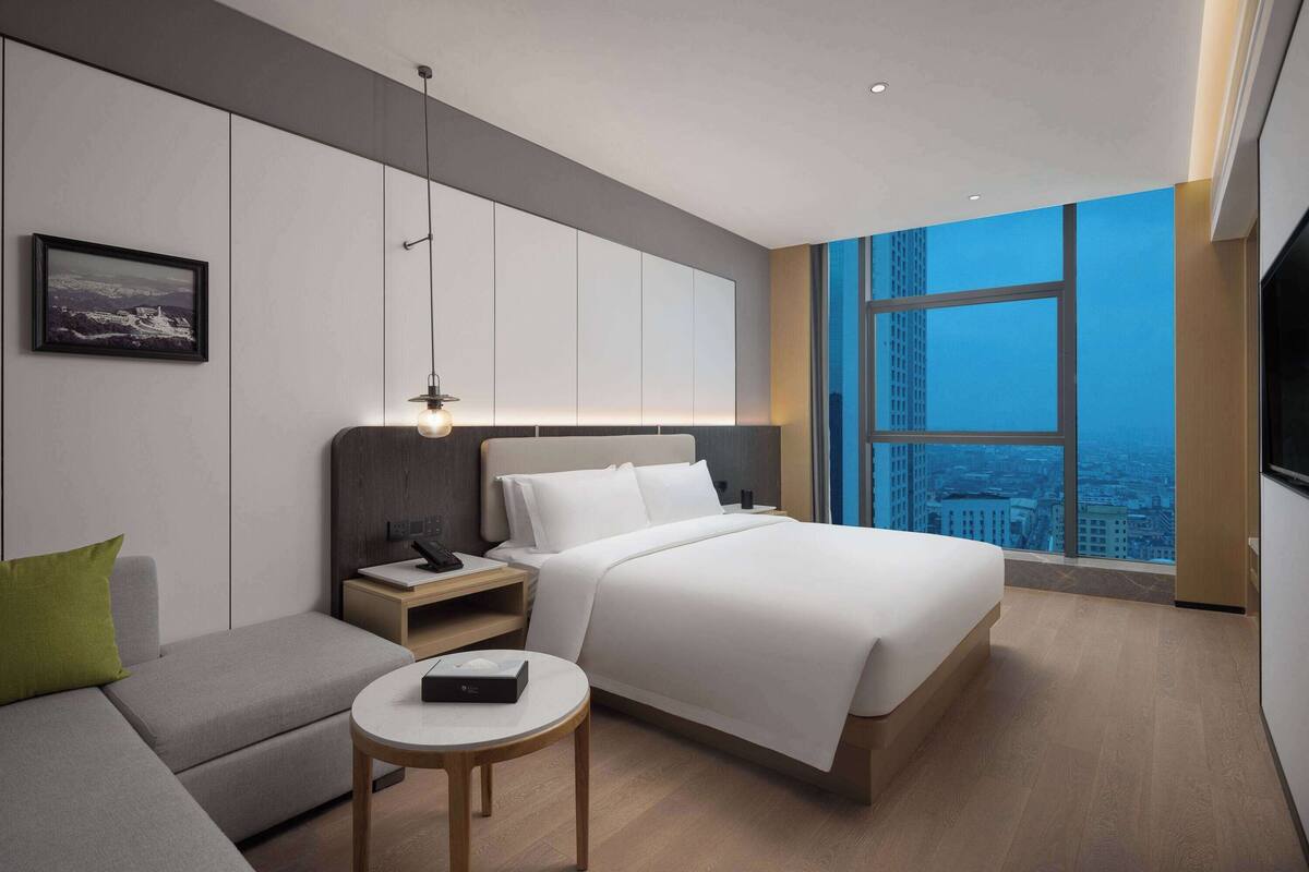A modern hotel room with a large bed, a sofa, a small table, and a city view through a large window.