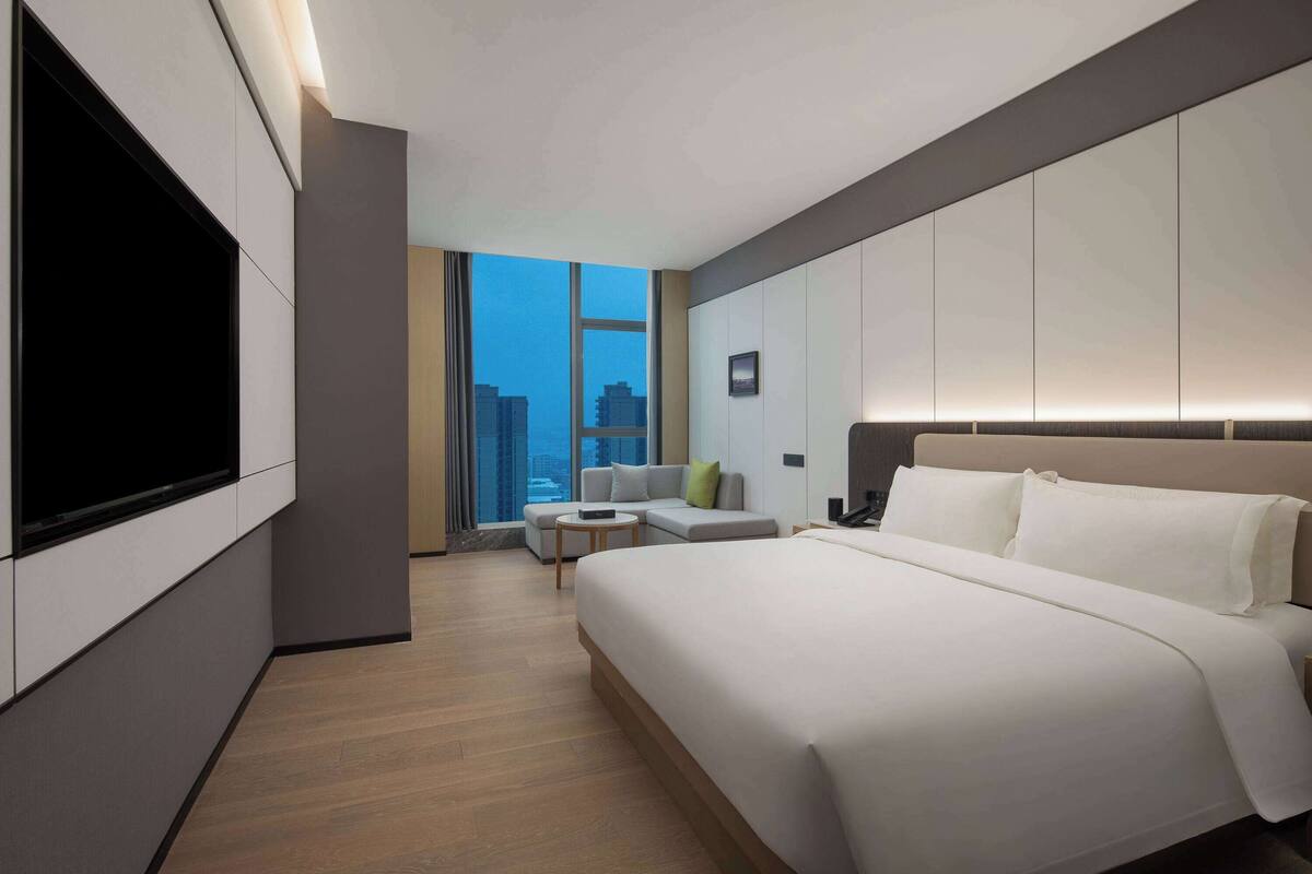 A modern hotel room with a large bed, a flat-screen TV mounted on the wall, a sofa, and a small table.