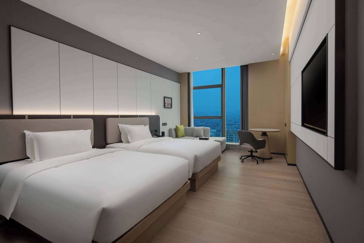 A modern hotel room with two beds, a desk, a chair, and a large window offering a city view.