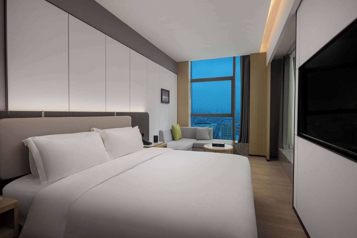 A modern hotel room with a large bed, a sofa, a small table, and a large window with a city view.