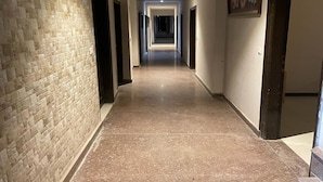 A long, narrow hallway with a stone wall on one side and a door on the other. The hallway has a polished floor and a ceiling with recessed lighting. - Hotel Pace Hero Honda Chowk (Gurugram)