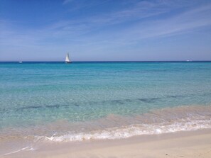 A beach with clear turquoise water, a sandy shore, and a sailboat in the distance.