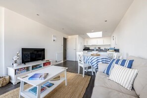 A modern living room with a flat-screen TV, a sofa, a coffee table, and a dining area with a striped tablecloth. - Modern Home with Sea Views, Newquay (Newquay)