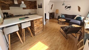 Une cuisine moderne avec un plan de travail blanc et des éléments en bois, un coin repas avec un canapé et une table basse, et un salon avec un canapé foncé et des coussins décoratifs.