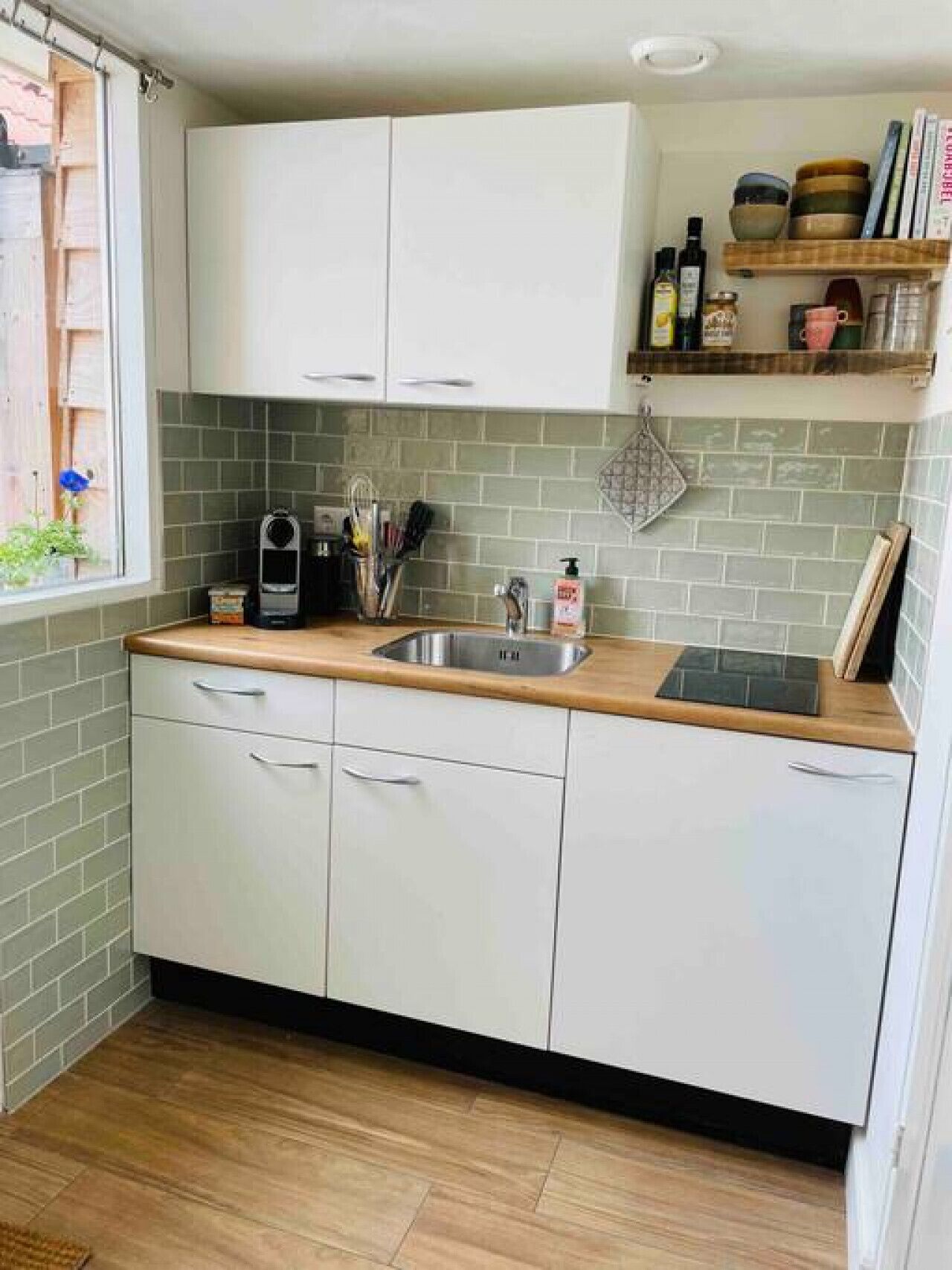 A compact kitchen with white cabinets, a wooden countertop, a stainless steel sink, and a window with a view of a garden.