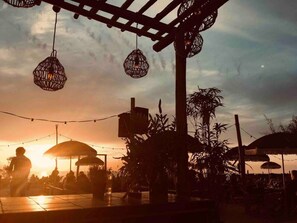 Un espacio al aire libre con mesas y sillas, una pérgola con luces colgantes y una puesta de sol al fondo.