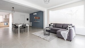 Un salon moderne avec un canapé gris, une table à manger et des chaises. Un tapis gris recouvre le sol, et un luminaire suspendu est fixé au plafond.