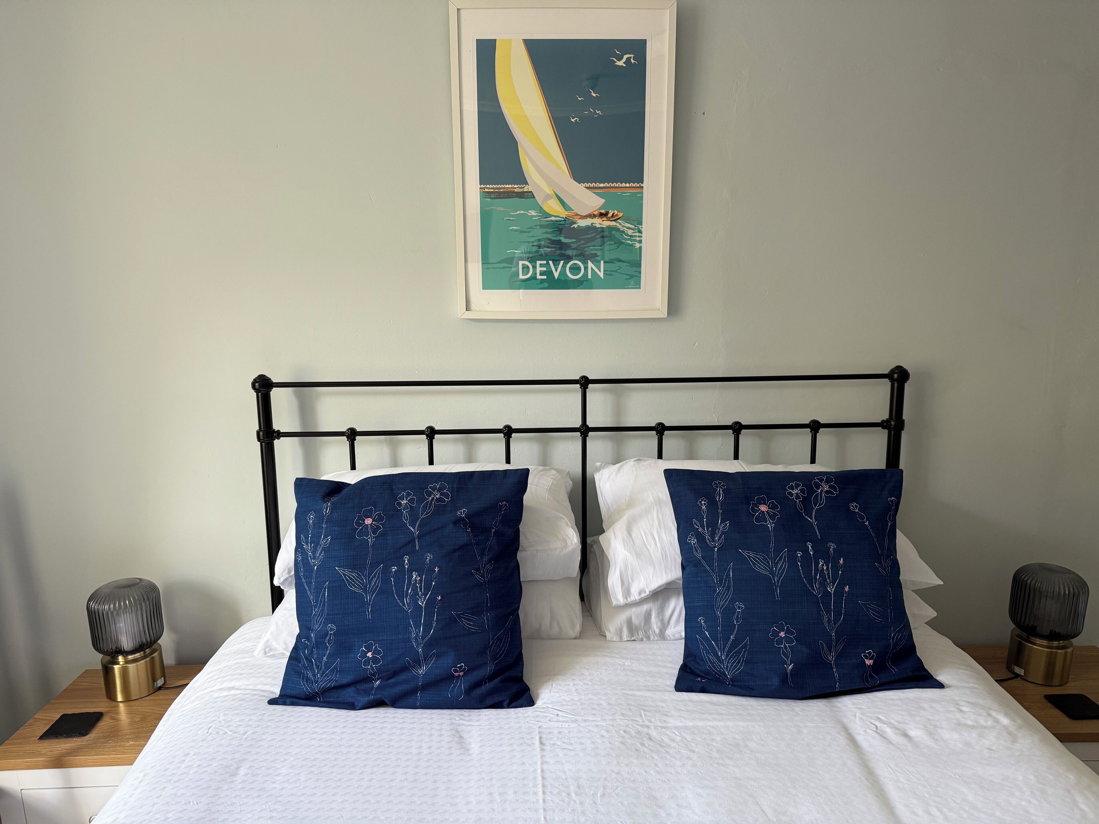 Ein Bett mit weißer Bettwäsche und zwei blaue Kissen mit Blumenmuster, ein gerahmtes Bild von einem Segelboot an der Wand und Nachttischlampen auf Holz-Nachttischen.