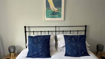 Ein Bett mit weißer Bettwäsche und zwei blaue Kissen mit Blumenmuster, ein gerahmtes Bild von einem Segelboot an der Wand und Nachttischlampen auf Holz-Nachttischen.