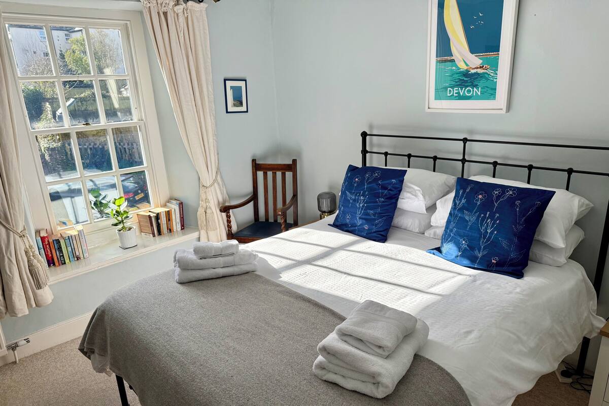 A neatly made bed with white and blue pillows, a grey blanket, and folded towels. There is a window with curtains, a chair, and a framed picture on the wall.