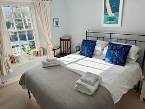 A neatly made bed with white and blue pillows, a grey blanket, and folded towels. There is a window with curtains, a chair, and a framed picture on the wall. - The Old Bakehouse (Colyton)