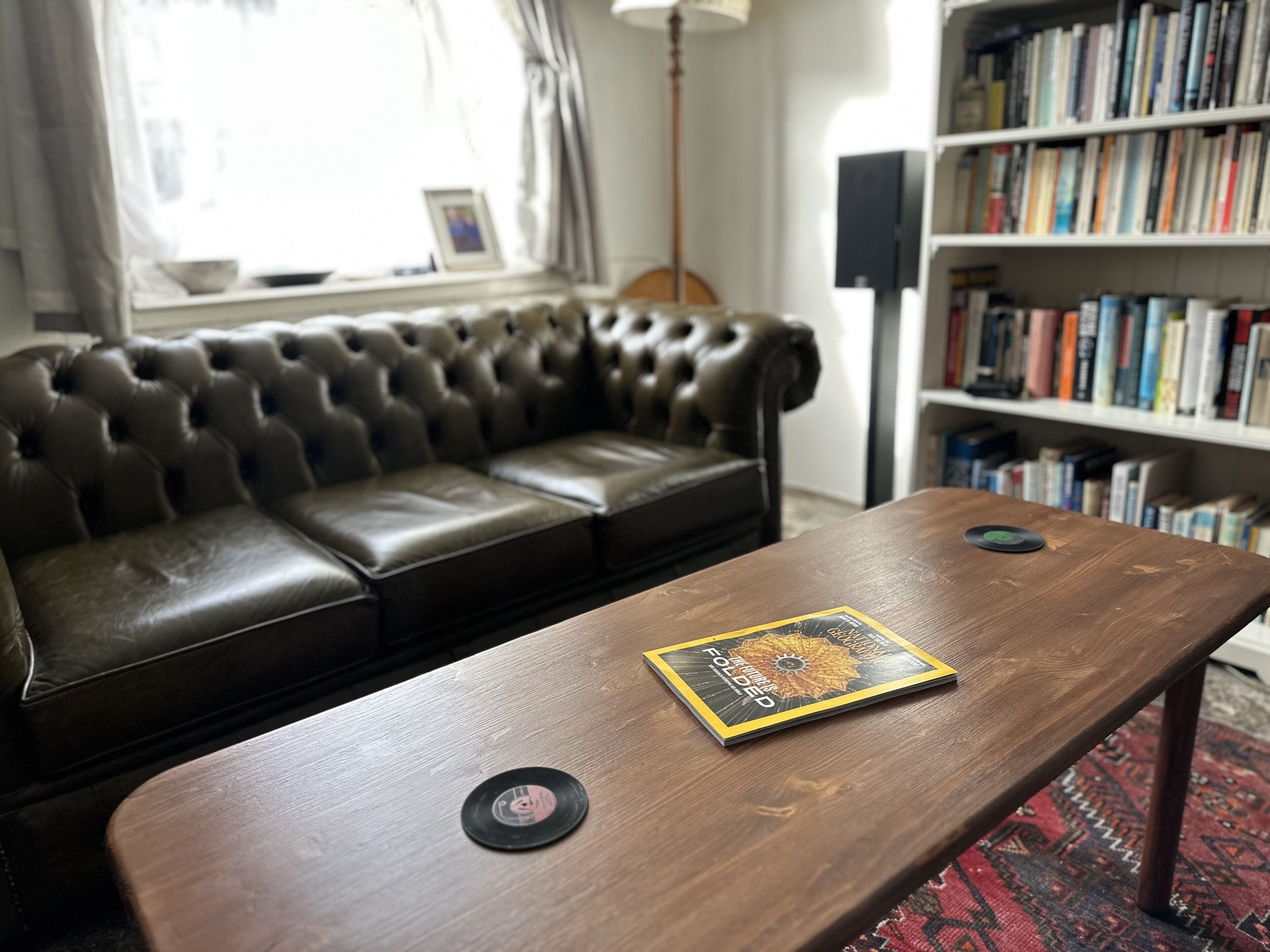 A room with a leather sofa, a wooden coffee table with vinyl records, a bookshelf, and a window with curtains.