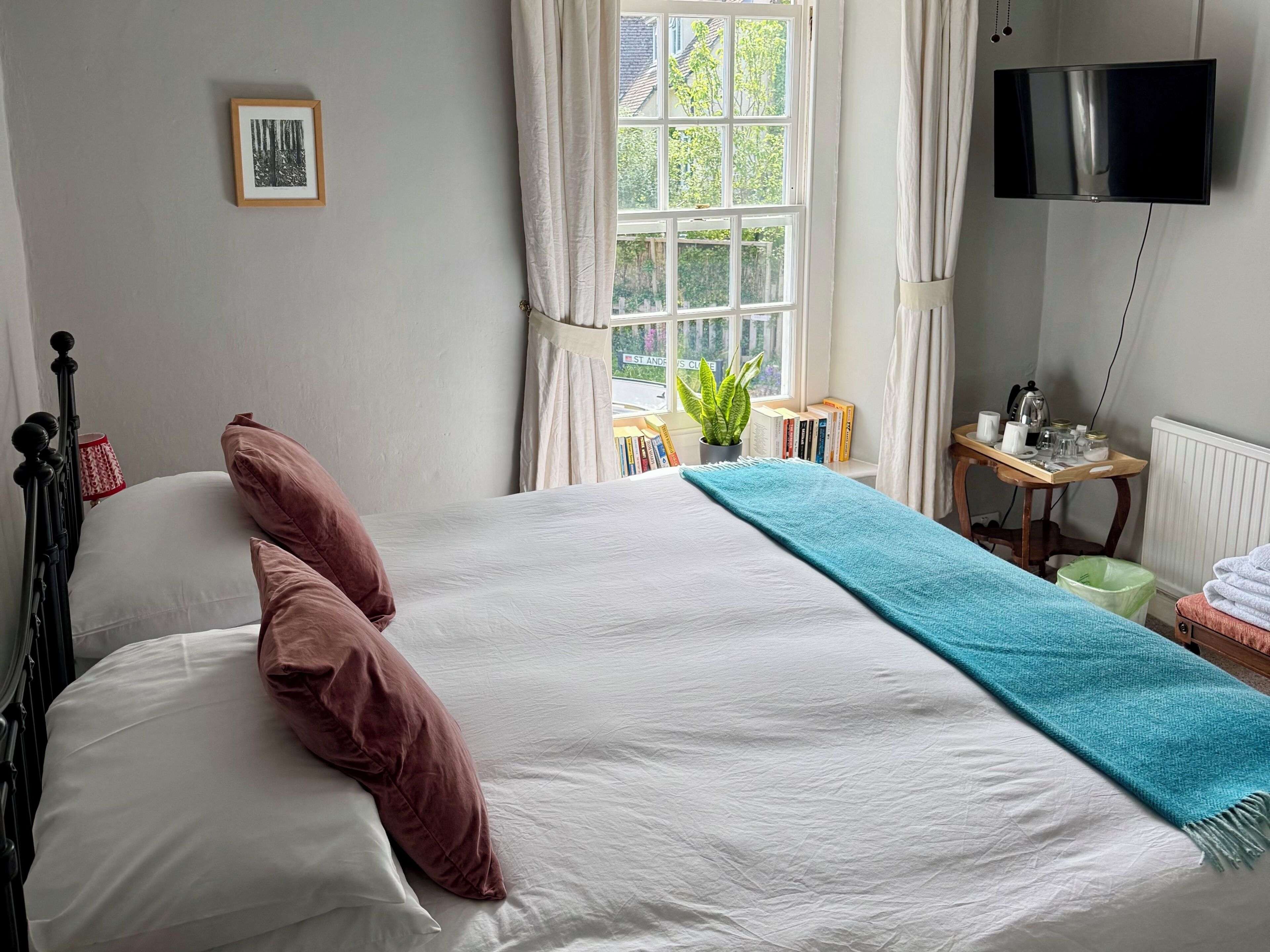 Un lit bien fait, agrémenté d’oreillers, une table de chevet avec des livres et une plante, une fenêtre avec des rideaux et un téléviseur à écran plat fixé au mur.