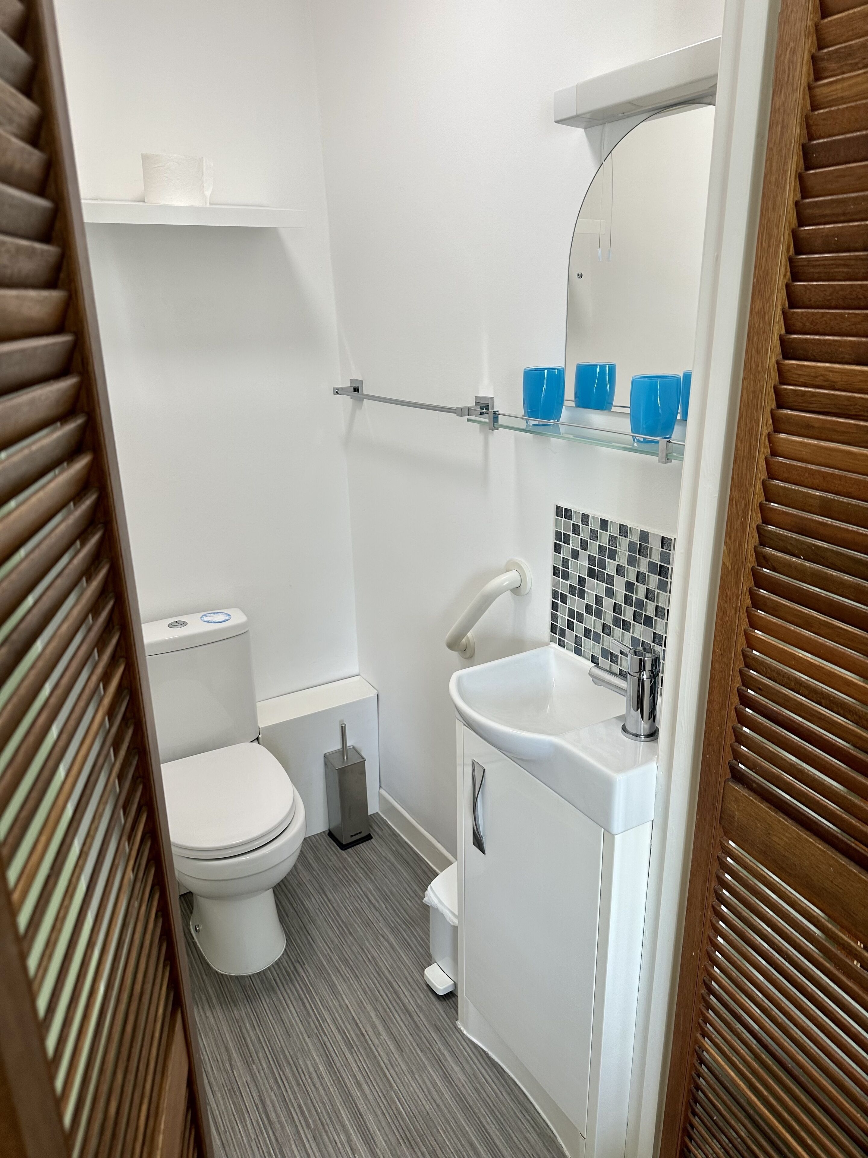 A compact bathroom with a toilet, a sink, and a mirror above it. There is a shelf with blue cups and a hanging towel rack.