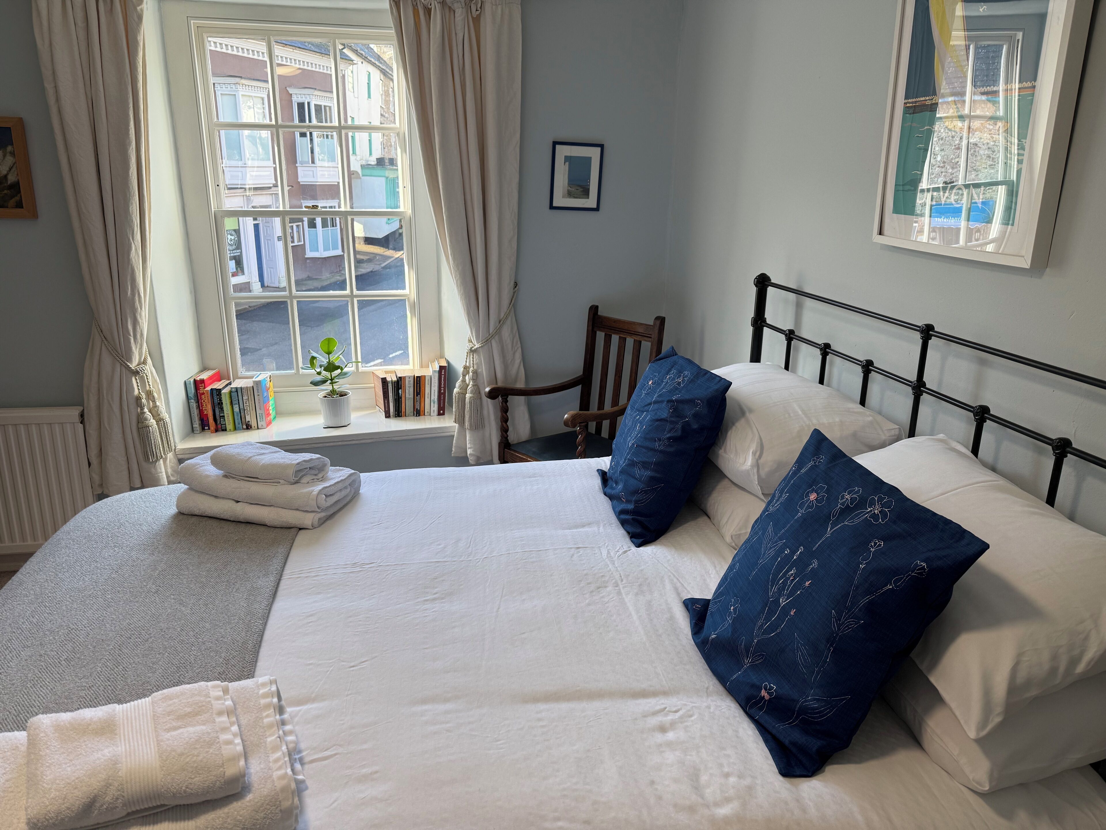 A neatly made bed with white and blue pillows, a bedside table with books, and a window with curtains.