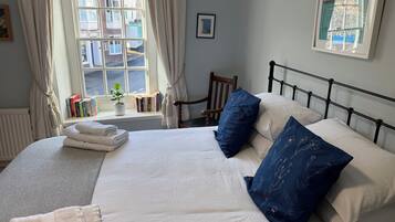A neatly made bed with white and blue pillows, a bedside table with books, and a window with curtains.