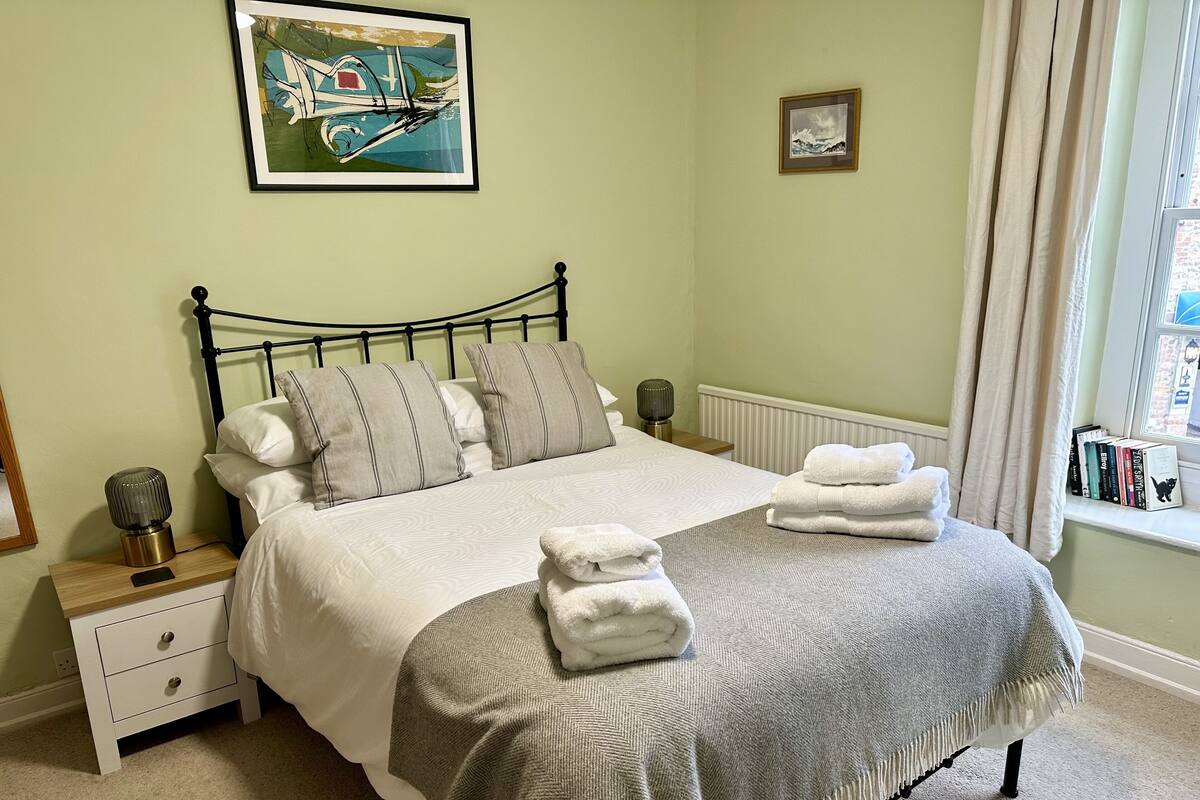 A neatly made bed with pillows, a bedside table with a lamp, and a window with books on the sill.
