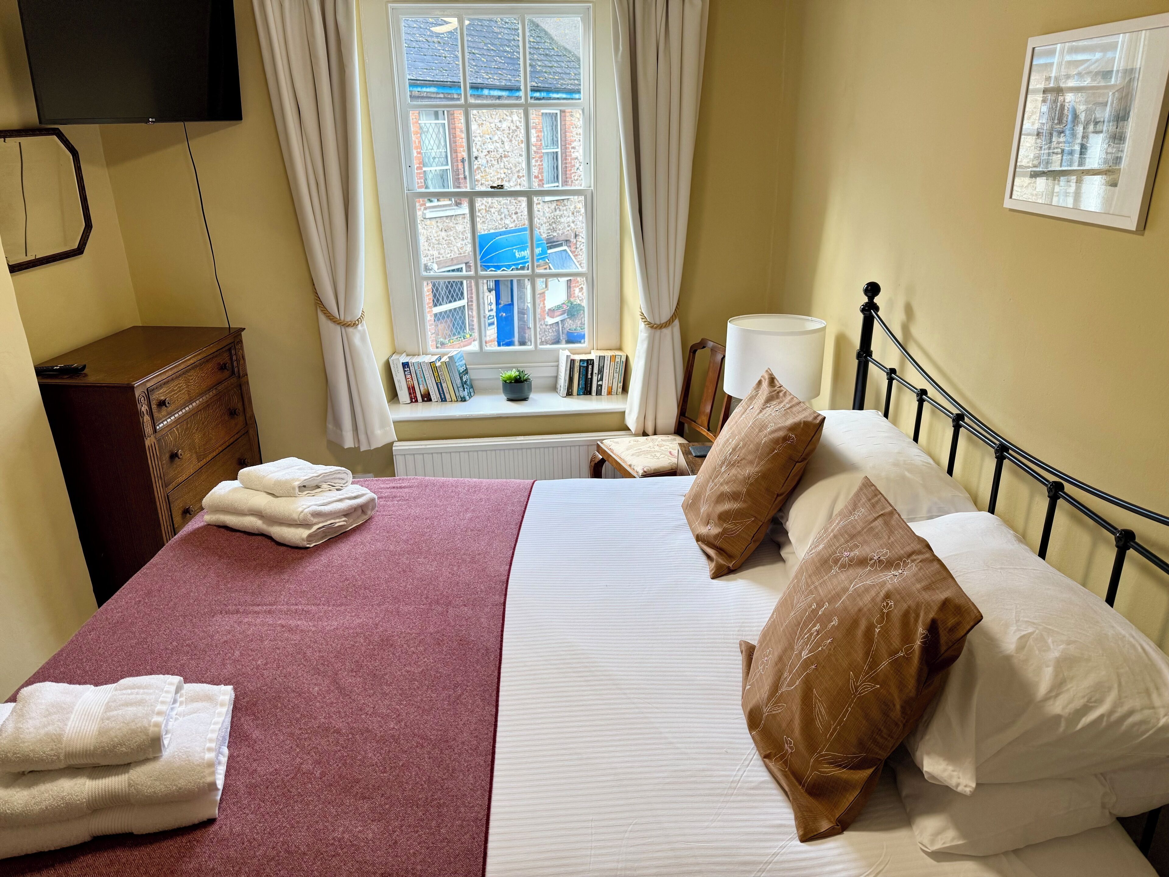A neatly made bed with pillows and folded towels, a bedside table with a lamp, a window with curtains, and a bookshelf with books.