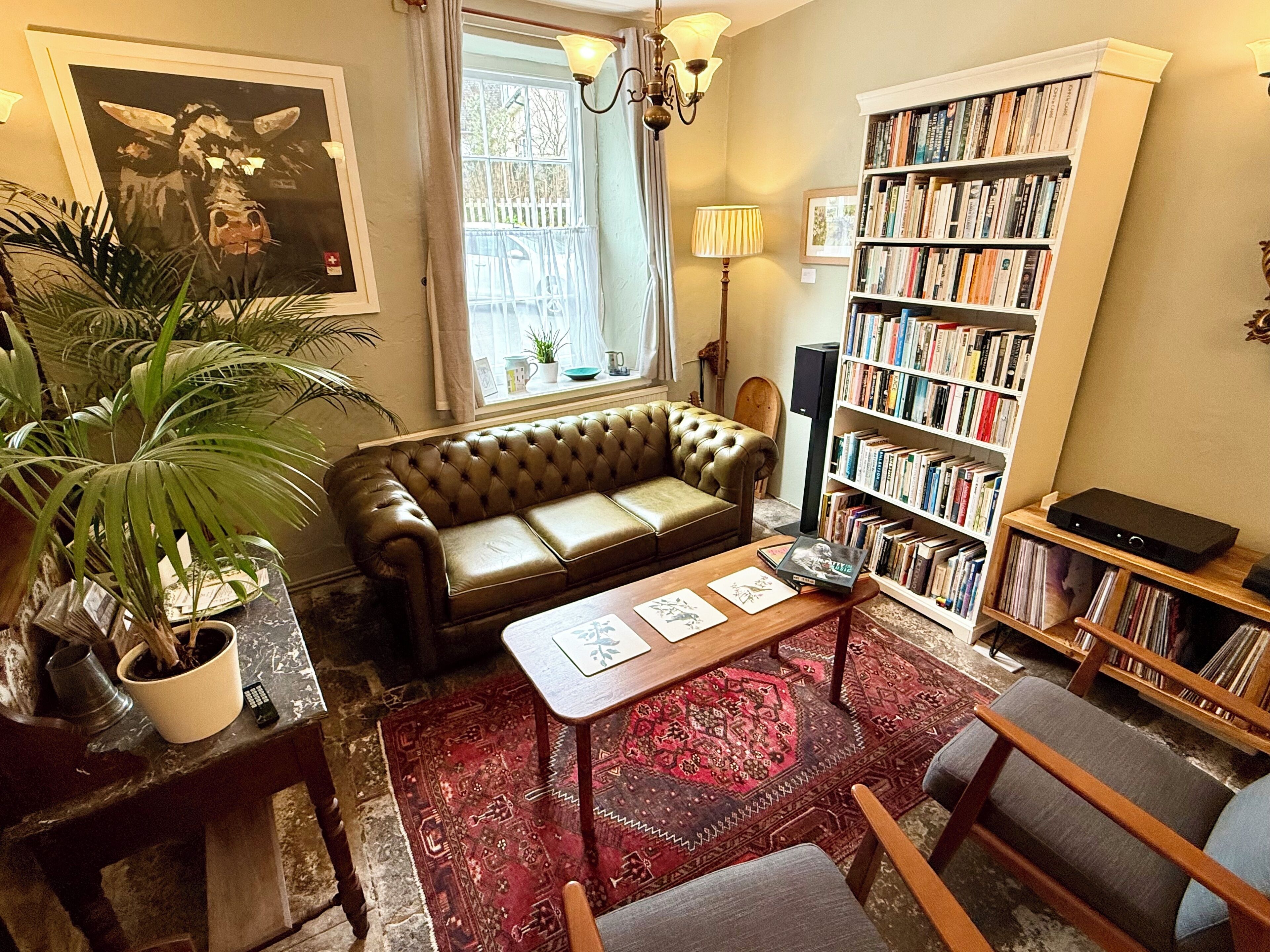Una sala de estar acogedora con un sofá de cuero marrón, una mesa de centro de madera, un tapete rojo estampado, una estantería llena de libros y una planta grande en maceta.