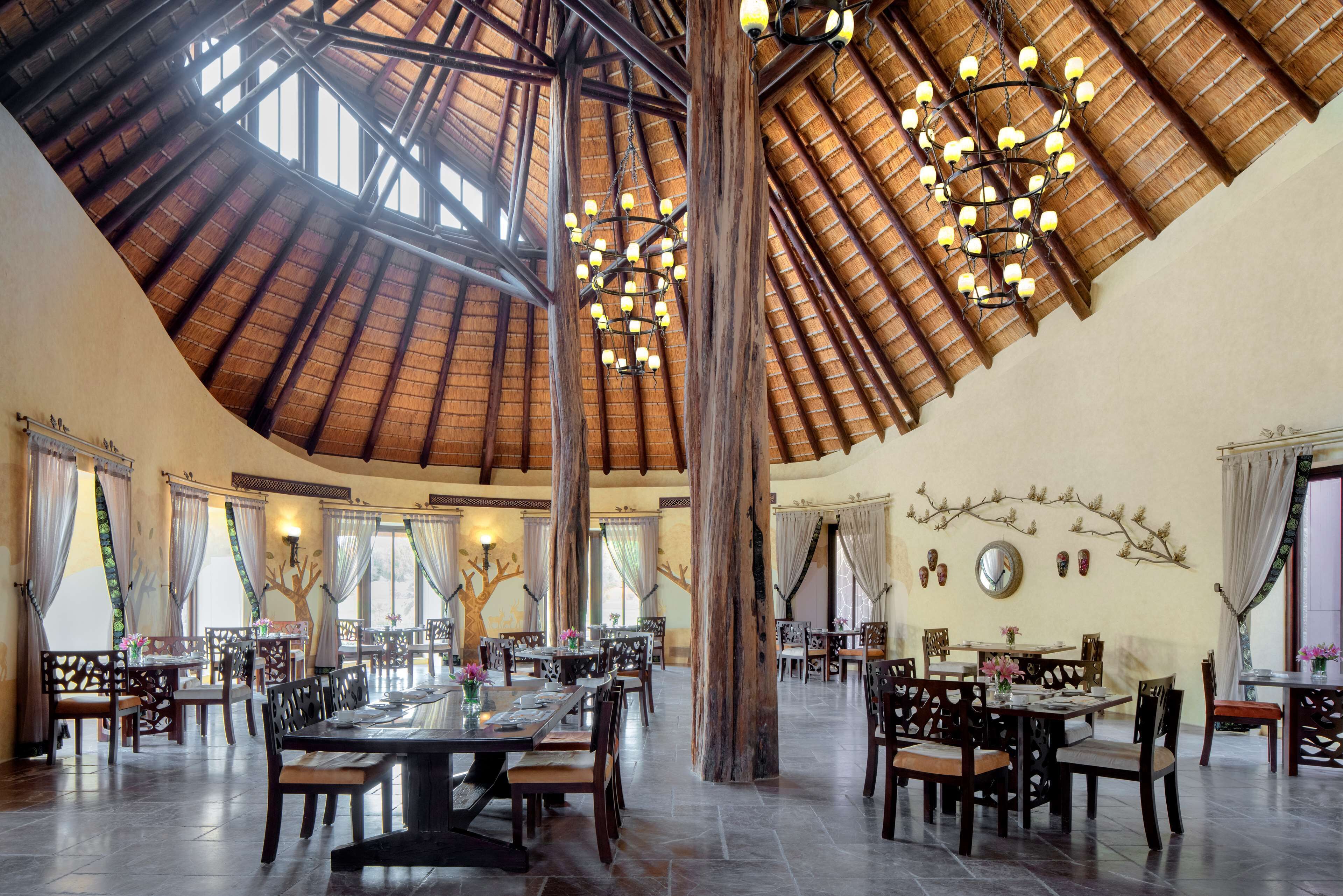 Uma sala de jantar ampla, com pé direito alto e vigas de madeira, apresentando várias mesas e cadeiras.