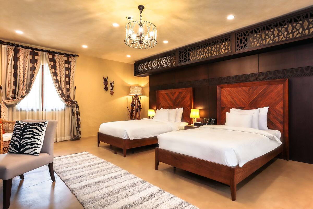 Habitación de hotel con dos camas, un candelabro, cabecera de madera, una lámpara y un cojín estampado con diseño de cebra.