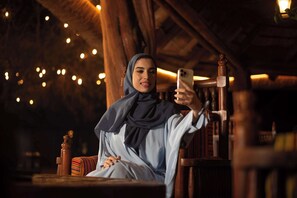 A person sitting in a wooden chair with a smartphone, in a room with wooden beams and string lights.