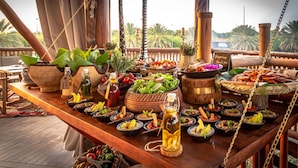 A wooden table with various dishes and a bottle of olive oil, set on a balcony with a view of palm trees.