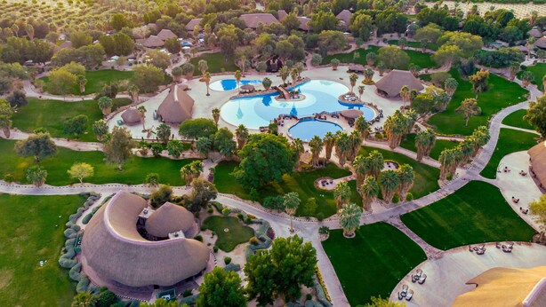 An aerial view of a resort with multiple swimming pools, palm trees, and thatched-roof structures.