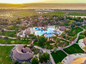 An aerial view of a resort with multiple swimming pools, palm trees, and thatched-roof structures.