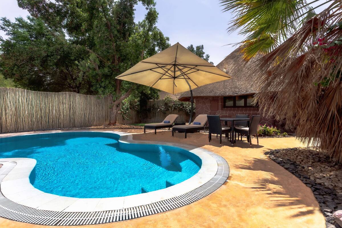Una piscina con borde curvo, rodeada por una terraza de concreto. Hay sillas de descanso y una mesa bajo un paraguas grande.