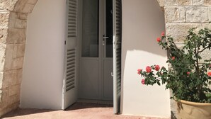 A stone archway leading to a door with a glass panel, a potted plant with red flowers, and a clear blue sky. - TRULLO SUITE 'THE QUEEN' in Itria Valley with swimming pool (Martina Franca)