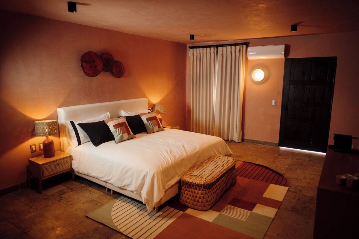 Un dormitorio con una cama grande, dos mesitas de noche con lámparas, un banco tejido y una alfombra estampada.