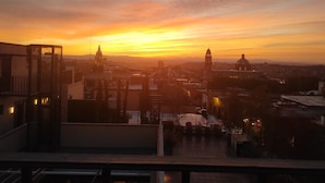 Vista del atardecer desde un balcón con vista a un paisaje urbano que incluye edificios y una torre de iglesia destacada.