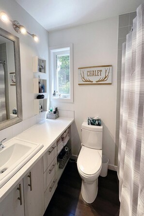 A modern bathroom with a large mirror, white vanity, and a toilet. There is a window providing natural light and a sign above the toilet. - Croissant des Soleils - RVMT (Mont-Tremblant)