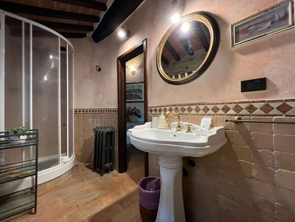 Un baño con lavamanos pedestal blanco, espejo redondo, radiador y cabina de ducha de vidrio.