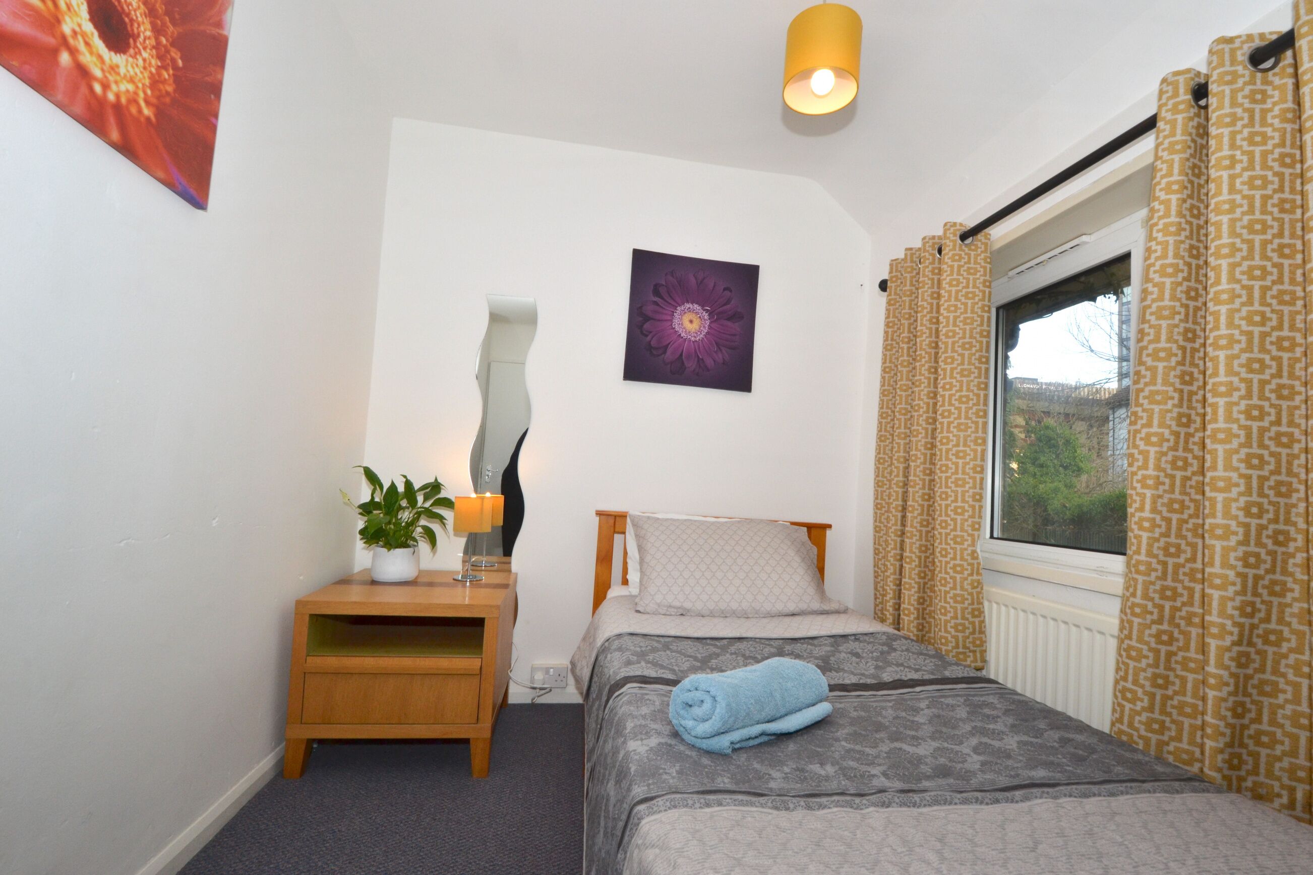 A bedroom with a bed, a bedside table with a lamp, a potted plant, and a window with curtains.