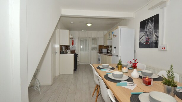 A modern kitchen with white cabinets, a dining area with a wooden table set for four, and a black and white cityscape painting on the wall. - Charming 5-bed House in City Center and Car Park (Birmingham)