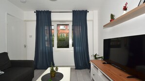 A modern living room with a black sofa, a coffee table with plants, a TV on a wooden stand, and a window with blue curtains. - Charming 5-bed House in City Center and Car Park (Birmingham)