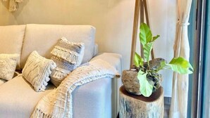 A modern living room with a sofa, decorative pillows, a macrame wall hanging, a lamp on a wooden stand, and a potted plant. - Beautiful apartment with pool (Sayulita)