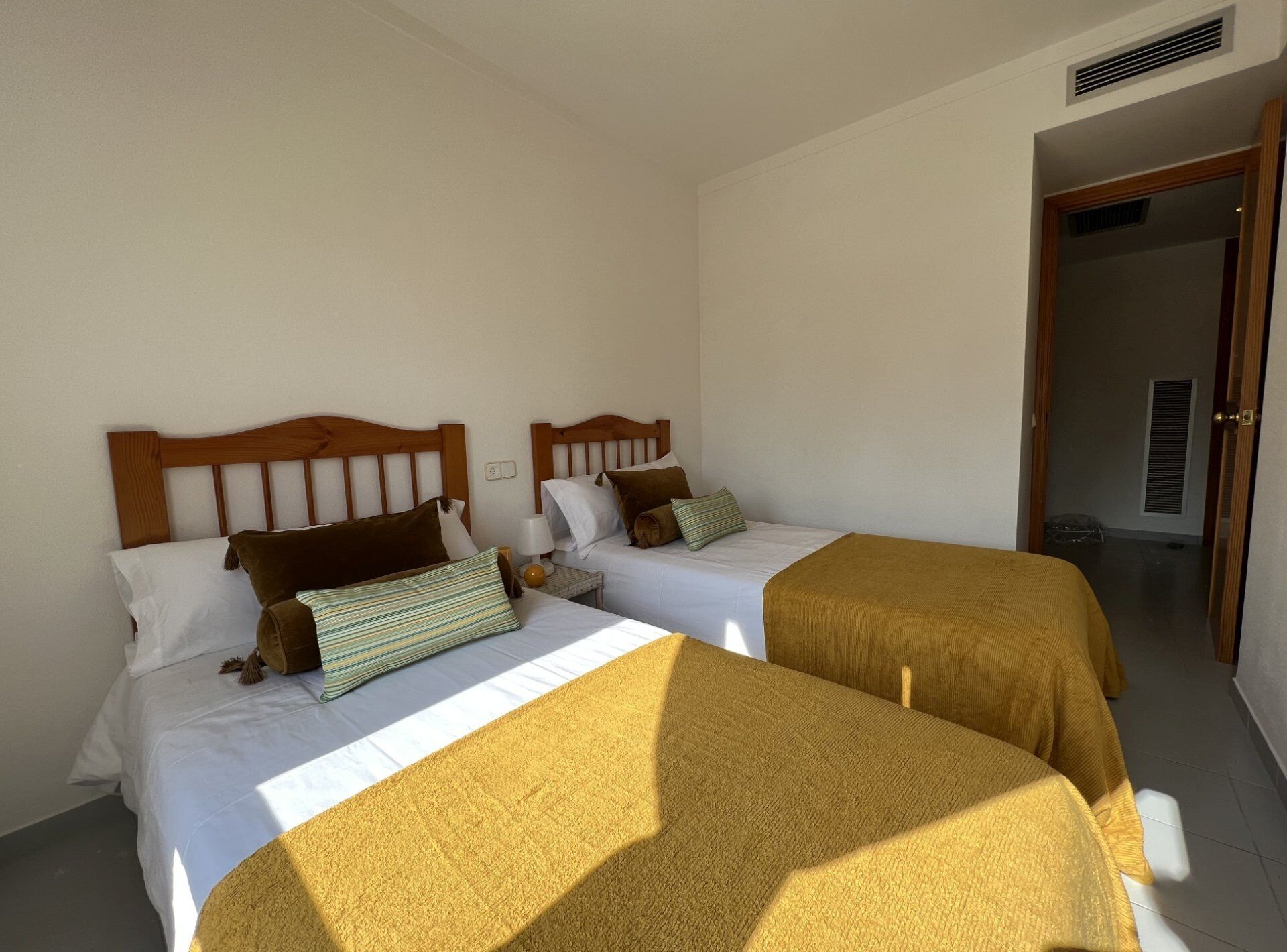 A hotel room with two beds, each with wooden headboards and white bedding. There is a door leading to another room and a vent on the wall.