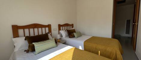 A hotel room with two beds, each with wooden headboards and white bedding. There is a door leading to another room and a vent on the wall.