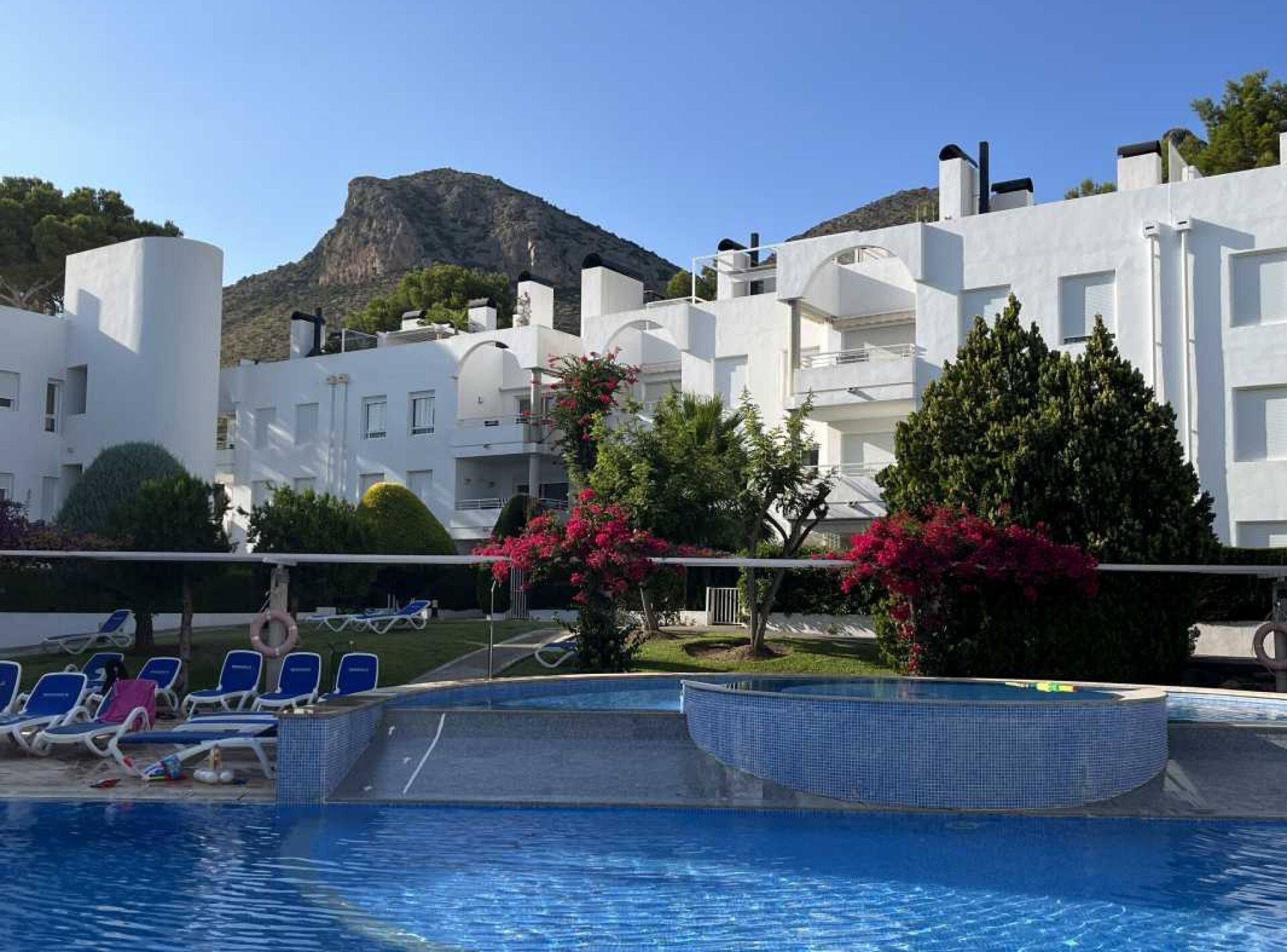 A swimming pool with lounge chairs, surrounded by white buildings with balconies, and a mountain in the background.