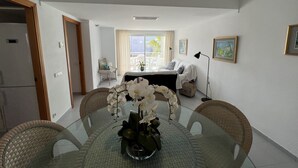 A dining area with a glass table surrounded by wicker chairs, a vase with white flowers, and a view of a balcony with a white sofa and a chair. - ISholidays Acron Puerto Pollensa (Port de Pollença)