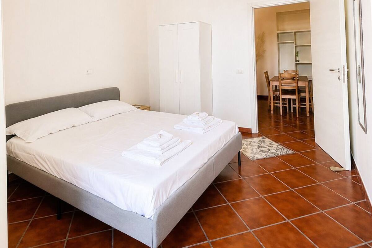 A bedroom with a bed, a wardrobe, a dining table with chairs, and a hanging light fixture.