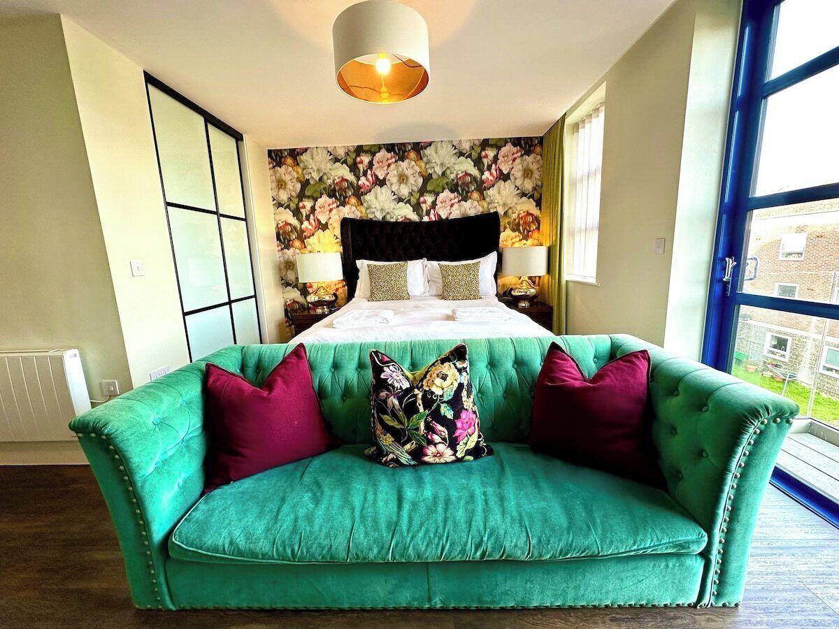 Ein Schlafzimmer mit einem grünen Sofa, einer blumigen Tagesdecke und Blick ins Freie durch ein großes Fenster.