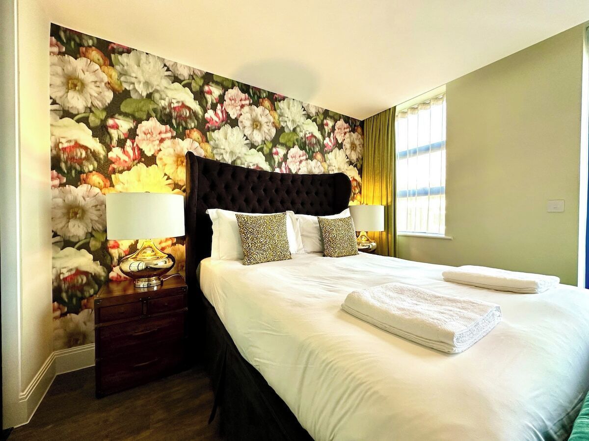 A hotel room with a large bed, floral wallpaper, a dark headboard, two bedside tables with lamps, and a window with curtains.