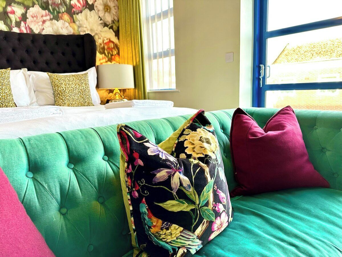 Una habitación con un sofá tapizado en verde, una cama con ropa blanca y una almohada estampada con motivos florales.